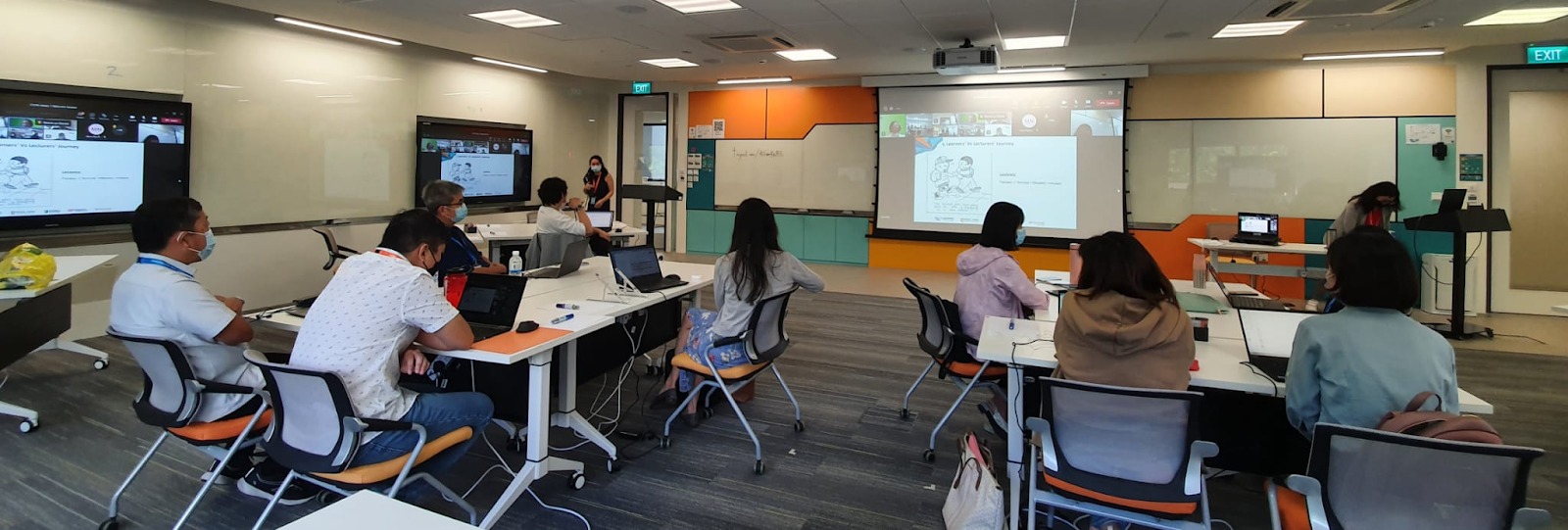 People seated at tables with laptops in a brightly lit classroom, facing a large projection screen and a presenter.